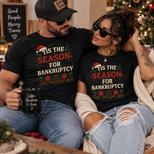 A man and a woman sit together in a cozy holiday setting, both wearing matching black graphic T-shirts that read 'Tis the Season for Bankruptcy' with a Santa hat and colorful Christmas floral design. The man holds a black mug that says 'Coffee, Christmas & Chaos'.