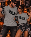A man and a woman pose together in a Halloween-themed setting, wearing matching heather gray graphic t-shirts with the text 'TAKE MY STRONG HAND!' and wing illustrations. The man holds a mug that says 'SCARY MOVIES BAD DECISIONS', and the woman holds a mug that says 'Just here for the Boo's'.