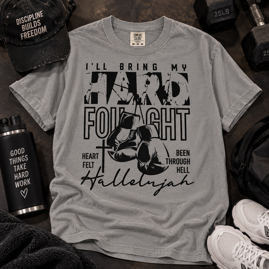 A light gray crewneck t-shirt with a black graphic print featuring boxing gloves, a cross, and motivational text, styled in a gym flat-lay with a black cap, water bottle, dumbbells, and sneakers.