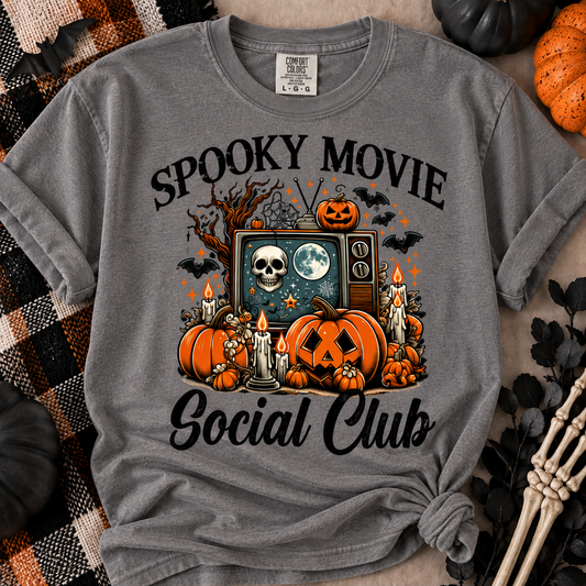 A heather gray graphic t-shirt featuring a Halloween-themed design with pumpkins, a TV showing a skull and moon, bats, candles, and the text 'SPOOKY MOVIE Social Club'. The shirt is styled in a flat-lay with autumn props including a skeleton hand, a plaid blanket, and pumpkins.