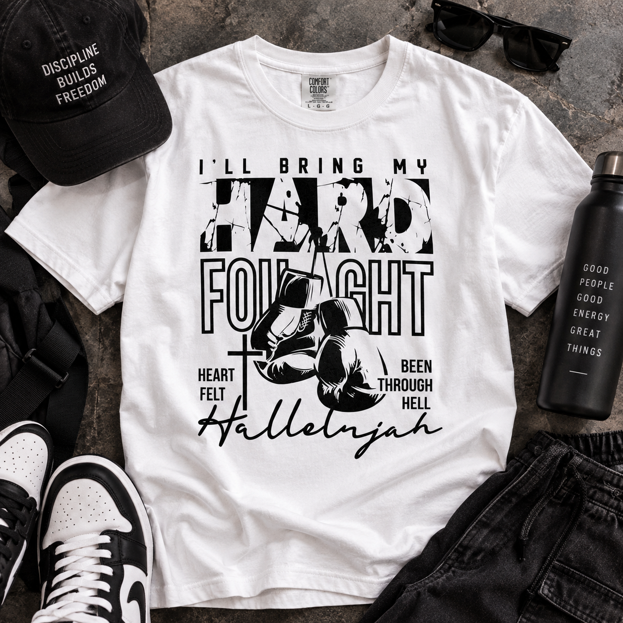 A white graphic t-shirt laid flat on a textured gray surface, surrounded by lifestyle props including a black baseball cap, sunglasses, a water bottle, sneakers, and black jeans. The t-shirt features a distressed black graphic of boxing gloves and the text 'I'LL BRING MY HARD FOILIGHT' and 'Hallelujah'.
