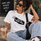 A woman sitting outdoors wearing a white graphic T-shirt with a black brushstroke design and the text 'TAKE MY STRONG HAND!' along with a hand gesture. She is also wearing ripped light-wash jeans, white sneakers, sunglasses, and carrying a black shoulder bag, while holding an iced coffee.
