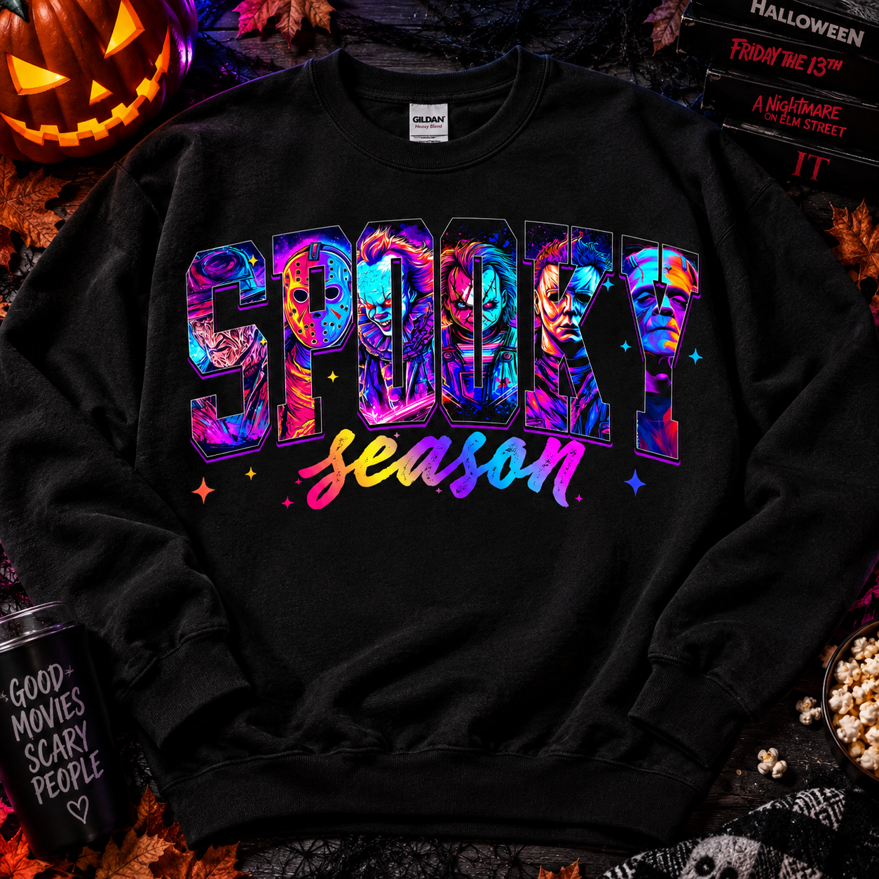 A black crewneck sweatshirt featuring a colorful graphic that reads 'SPOOKY season' with horror character faces inside the letters. The sweatshirt is displayed in a flat-lay Halloween setting with a jack-o'-lantern, popcorn, and themed books.