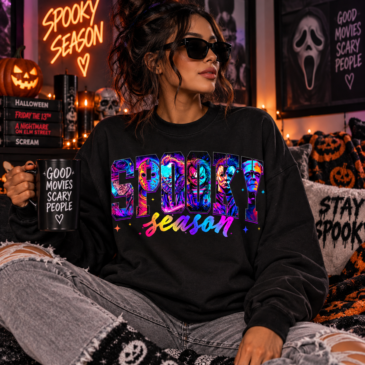 A woman wearing a black sweatshirt with a colorful 'SPOOKY season' graphic sits in a cozy, Halloween-themed room. She holds a black mug with the text 'GOOD MOVIES SCARY PEOPLE' and wears ripped gray jeans.