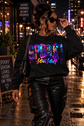 A woman walking on a wet city street at night wearing a black crewneck sweatshirt with a colorful 'Spooky Season' graphic print, paired with black leather pants and sunglasses.