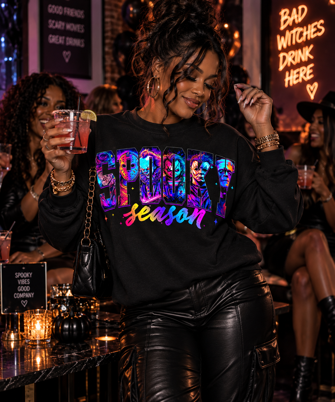 A woman in a black leather-look pants and a black sweatshirt with a colorful 'Spooky Season' graphic stands in a dimly lit bar holding a drink. The background features neon signs and other patrons.