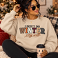A woman wearing a cream-colored crewneck sweatshirt with the text 'I DON'T DO WINTER very well' sits on a cozy couch holding a coffee cup, with a decorated Christmas tree in the background.