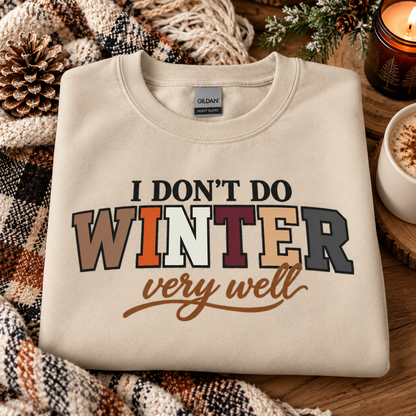 A folded cream-colored Gildan sweatshirt with a winter-themed graphic reading 'I DON'T DO WINTER very well' in multicolor lettering, styled on a rustic wooden surface with a plaid blanket, pinecone, and a lit candle.