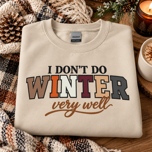 A folded cream-colored Gildan sweatshirt with a winter-themed graphic reading 'I DON'T DO WINTER very well' in multicolor lettering, styled on a rustic wooden surface with a plaid blanket, pinecone, and a lit candle.