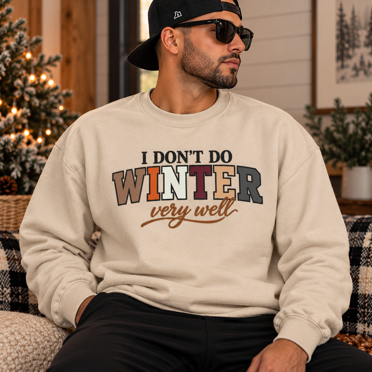 A man wearing a beige crewneck sweatshirt with the text 'I DON'T DO WINTER very well' in a multicolor block-letter design, sitting in a cozy indoor setting with a Christmas tree in the background.
