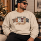 A man wearing a beige crewneck sweatshirt with the text 'I DON'T DO WINTER very well' in a multicolor block-letter design, sitting in a cozy indoor setting with a Christmas tree in the background.