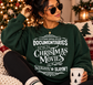 A woman wearing a dark green crewneck sweatshirt with a white graphic text print that reads 'IT'S EITHER SERIAL KILLER DOCUMENTARIES OR CHRISTMAS MOVIES OR OTHER SLEIGHIN' & SLAYIN'!', sitting in a cozy holiday setting with a Christmas tree and warm lights in the background.