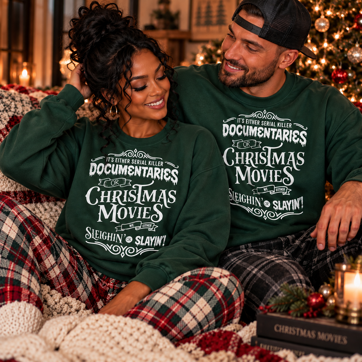 A couple sitting together on a couch in a cozy holiday setting, wearing matching dark green crewneck sweatshirts with white text that reads 'IT'S EITHER ROBIN KILLER DOCUMENTARIES CHRISTMAS MOVIES SLEIGHIN' SLAYIN!'.