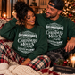 A couple sitting together on a couch in a cozy holiday setting, wearing matching dark green crewneck sweatshirts with white text that reads 'IT'S EITHER ROBIN KILLER DOCUMENTARIES CHRISTMAS MOVIES SLEIGHIN' SLAYIN!'.