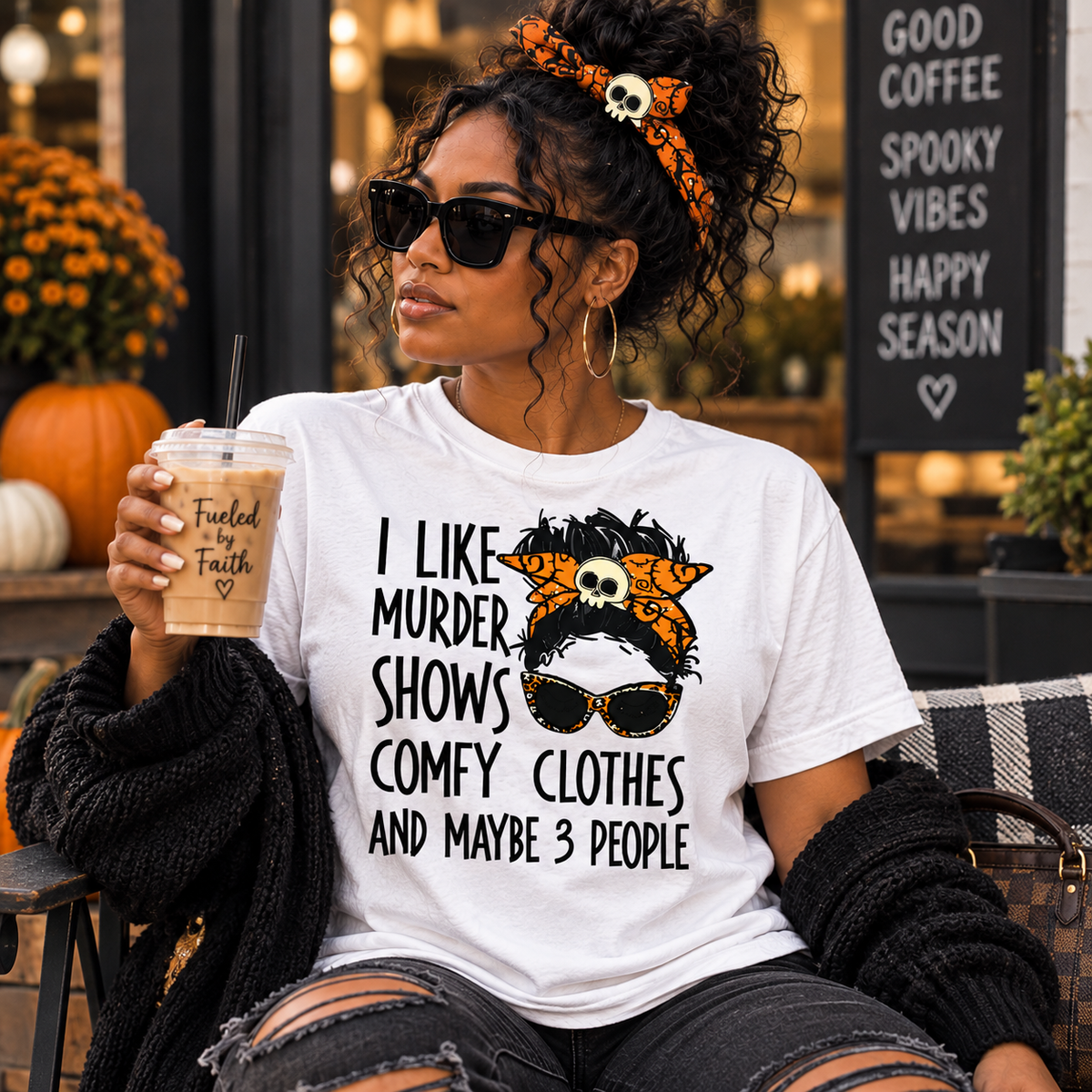 A woman wearing a white graphic t-shirt with a horror-comedy design that reads 'I LIKE MURDER SHOWS COMFY CLOTHES AND MAYBE 3 PEOPLE' next to a skull-and-orange-pattern headband, holding an iced coffee in a fall-themed outdoor setting.