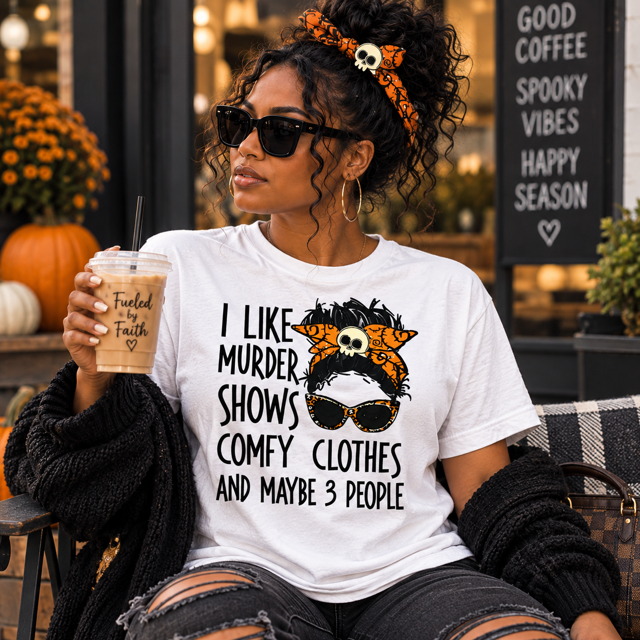 A woman wearing a white graphic t-shirt with a horror-comedy design that reads 'I LIKE MURDER SHOWS COMFY CLOTHES AND MAYBE 3 PEOPLE' next to a skull-and-orange-pattern headband, holding an iced coffee in a fall-themed outdoor setting.