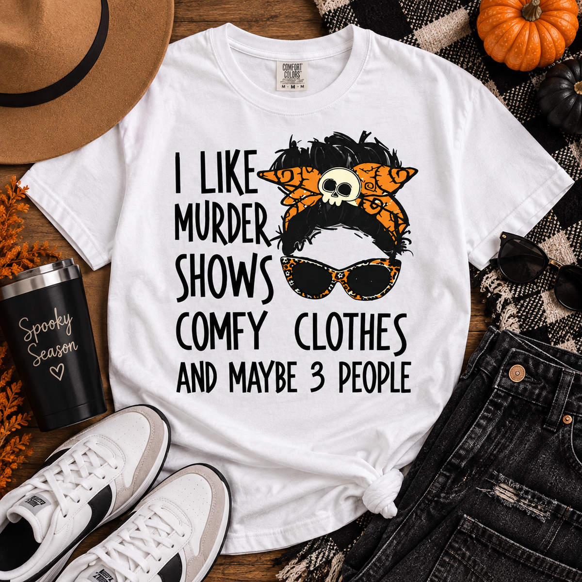 A white graphic T-shirt laid flat on a wood surface with styled seasonal props including a tan hat, black tumbler, white sneakers, dark jeans, sunglasses, pumpkins, and a plaid blanket. The shirt has black text reading 'I LIKE MURDER SHOWS COMFY CLOTHES AND MAYBE 3 PEOPLE' with a stylized black-and-orange hair accessory illustration and skull detail.