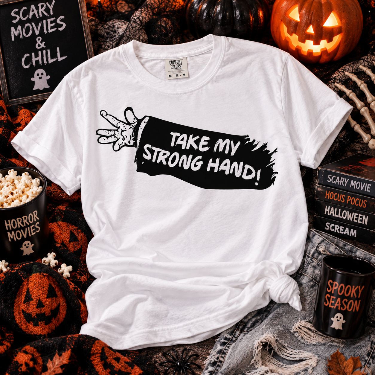 A white short-sleeve graphic T-shirt laid flat in a Halloween-themed setting, with a black banner-style print reading “TAKE MY STRONG HAND!” and a hand illustration. The shirt is surrounded by pumpkins, popcorn, books, and mugs with horror movie text.