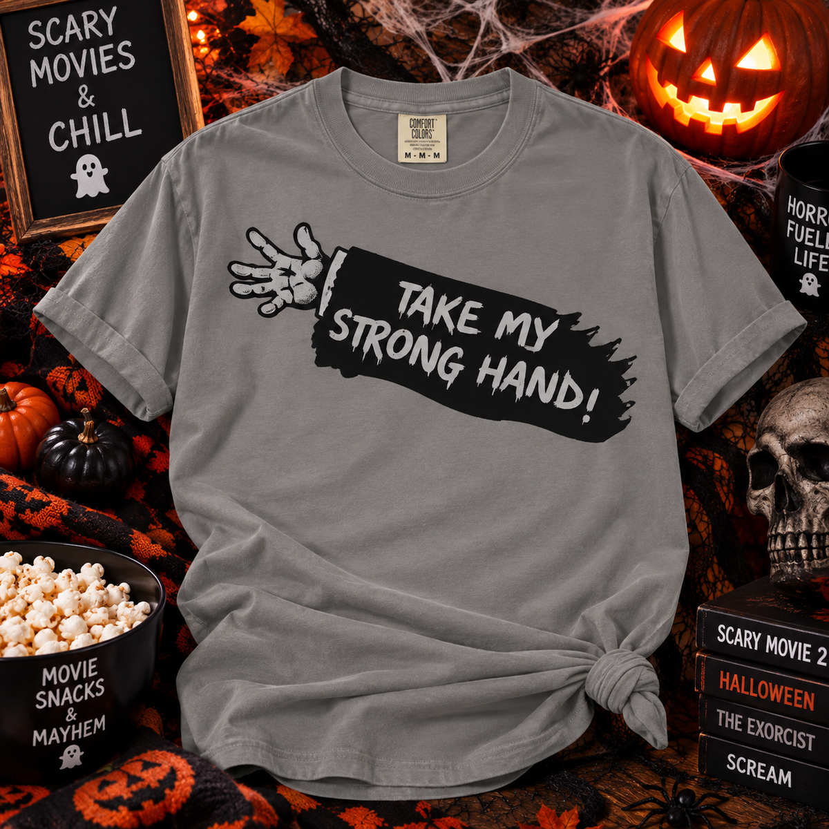 A heather gray graphic t-shirt with a distressed black paint stroke design and a skeleton hand graphic, featuring the text 'TAKE MY STRONG HAND!'. The shirt is styled in a Halloween-themed flat-lay setting with pumpkins, a skull, popcorn, and horror movie props.