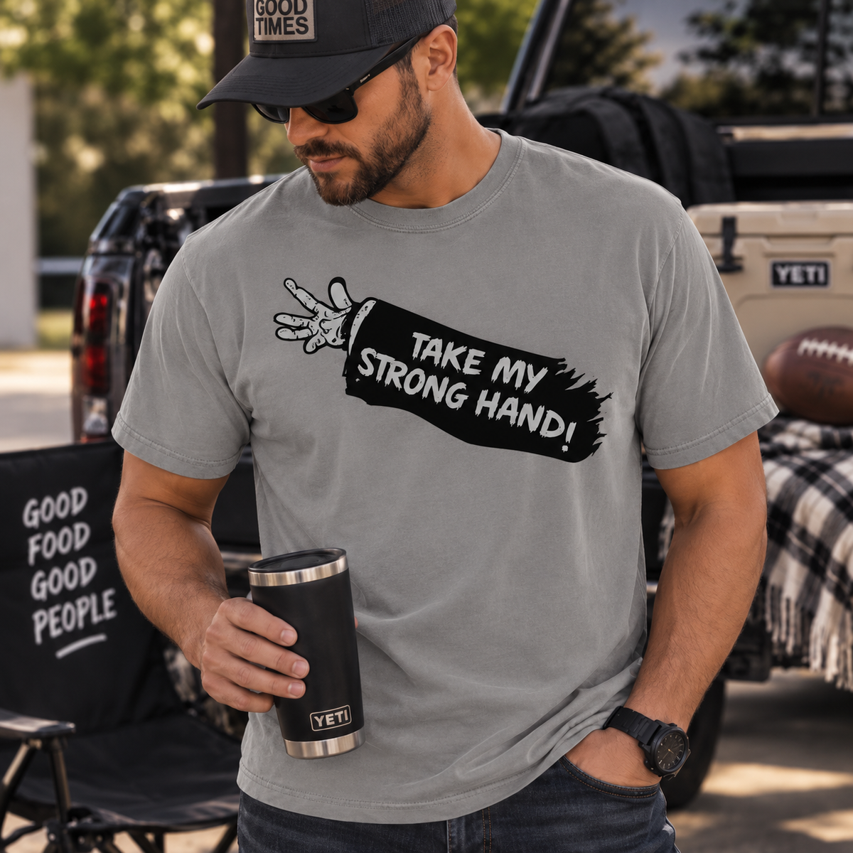 A man wearing a gray t-shirt with a black graphic that reads 'TAKE MY STRONG HAND!' and holding a black YETI tumbler, set against an outdoor tailgate background with a YETI cooler and truck.