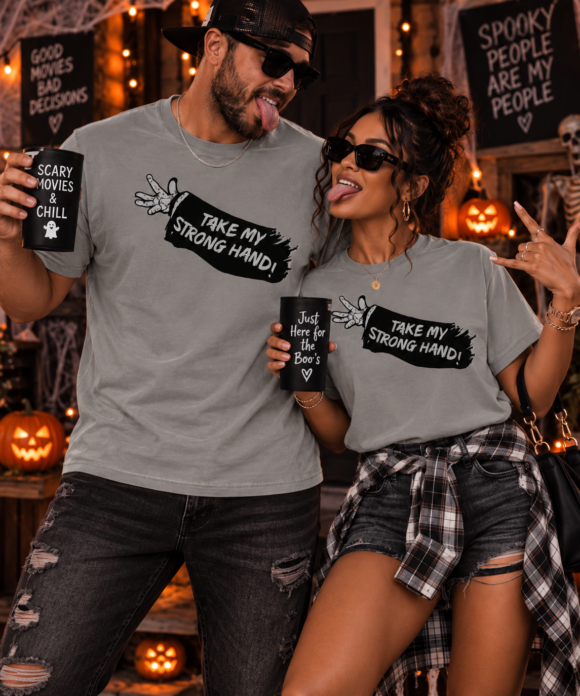 A man and a woman pose together in a Halloween-themed setting, wearing matching heather gray graphic t-shirts with the text 'TAKE MY STRONG HAND!' and wing illustrations. The man holds a mug that says 'SCARY MOVIES BAD DECISIONS', and the woman holds a mug that says 'Just here for the Boo's'.