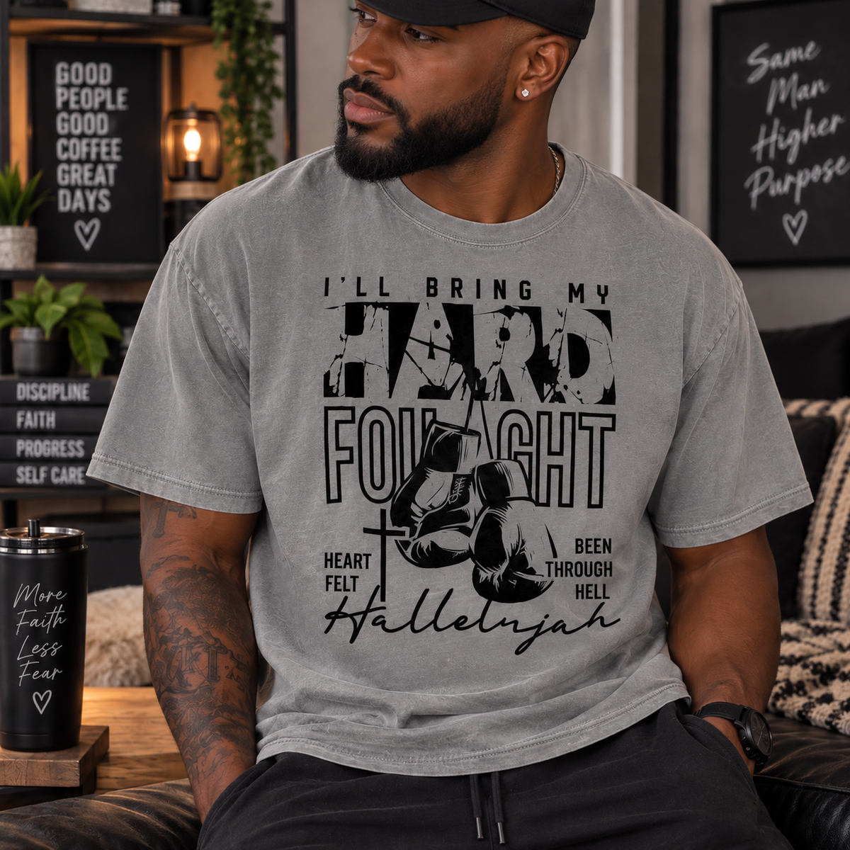 A man wearing a heather gray graphic t-shirt with a black print that reads 'I'LL BRING MY HARD FOYIGHT' and 'Hallelujah' with a cross and boxing gloves graphic. The setting is a cozy, dimly lit living room with motivational posters in the background.