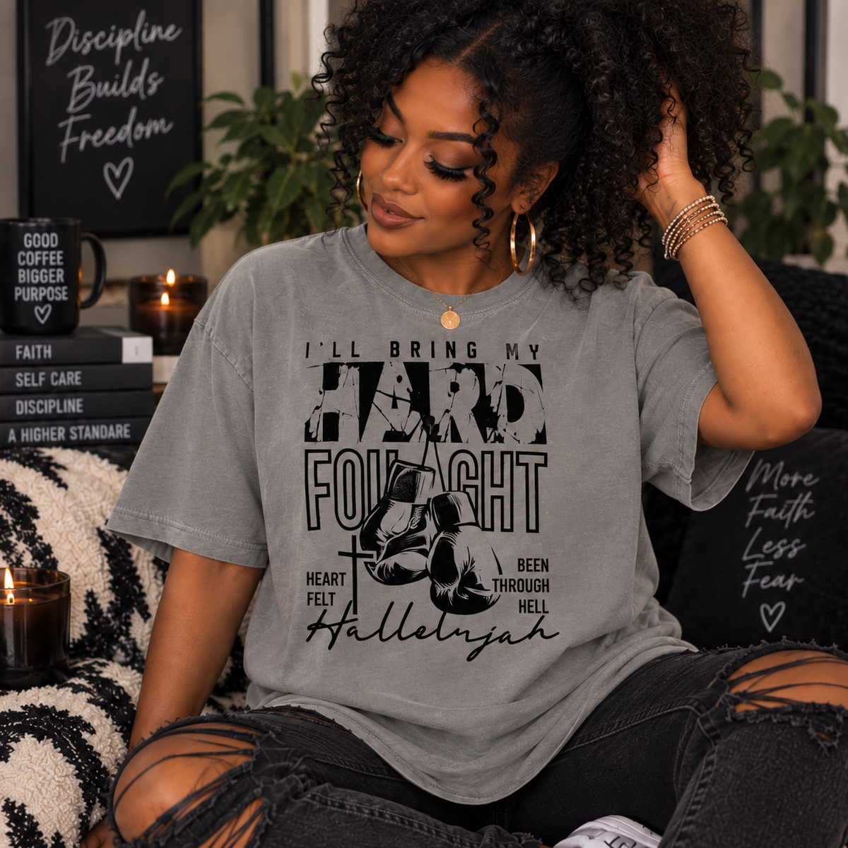 A woman wearing a heather gray graphic t-shirt with black text and a cross design that reads 'I'LL BRING MY HARD FIGHT', sitting in a cozy, dimly lit room with candles and books.