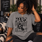 A woman wearing a heather gray graphic t-shirt with black text and a cross design that reads 'I'LL BRING MY HARD FIGHT', sitting in a cozy, dimly lit room with candles and books.