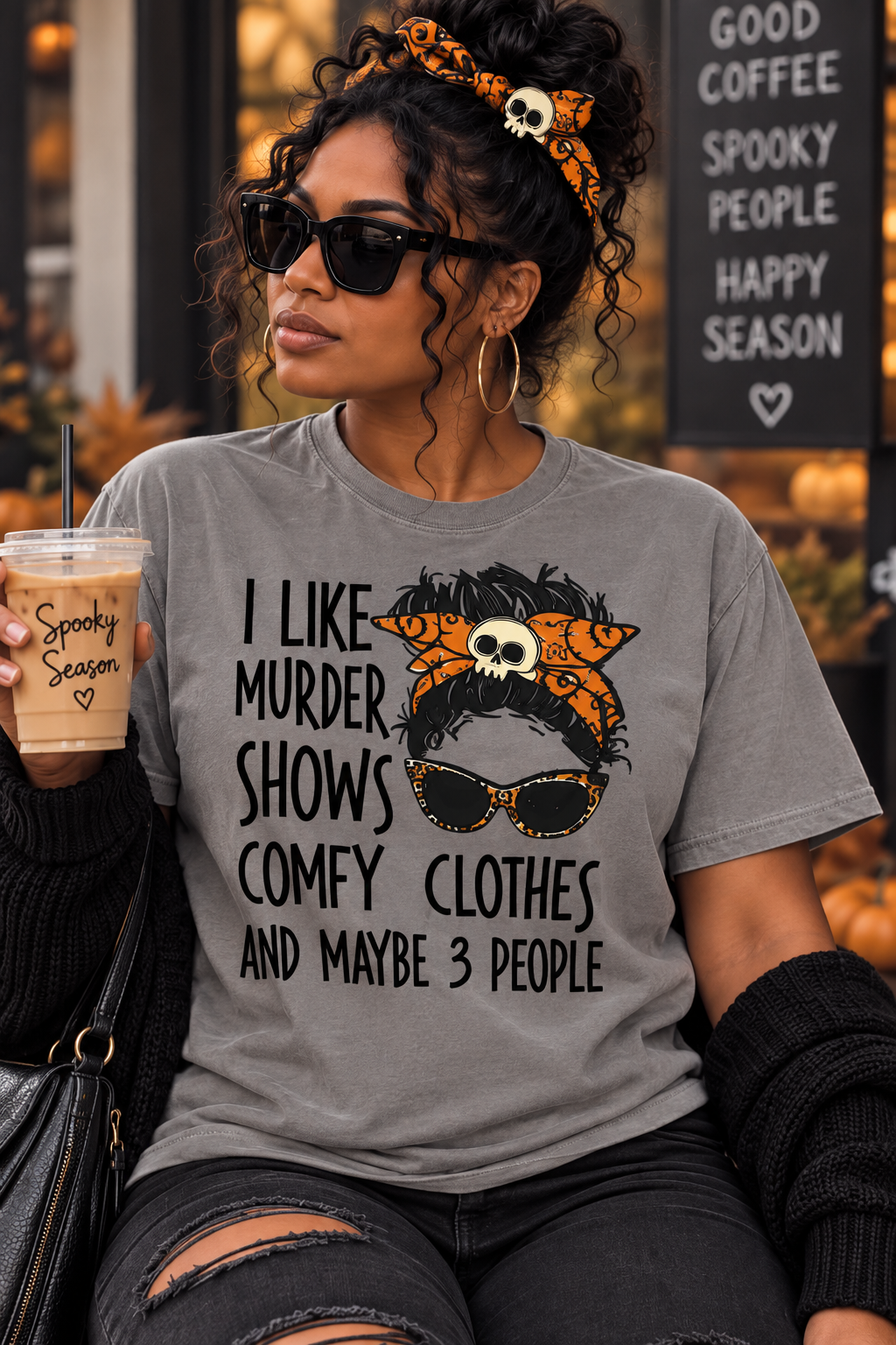 A woman wearing a heather gray graphic t-shirt with a Halloween-themed design. The graphic features a woman with a skull-patterned headband, black sunglasses, and orange-and-black cat ears, accompanied by the text 'I LIKE MURDER SHOWS COMFY CLOTHES AND MAYBE 3 PEOPLE'.
