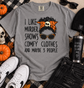 A heather gray graphic t-shirt laid flat on a plaid blanket with autumn props. The shirt features a black text graphic reading 'I LIKE MURDER SHOWS COMFY CLOTHES AND MAYBE 3 PEOPLE' alongside an illustration of a woman with a skull-and-orange headwrap and leopard-print sunglasses.